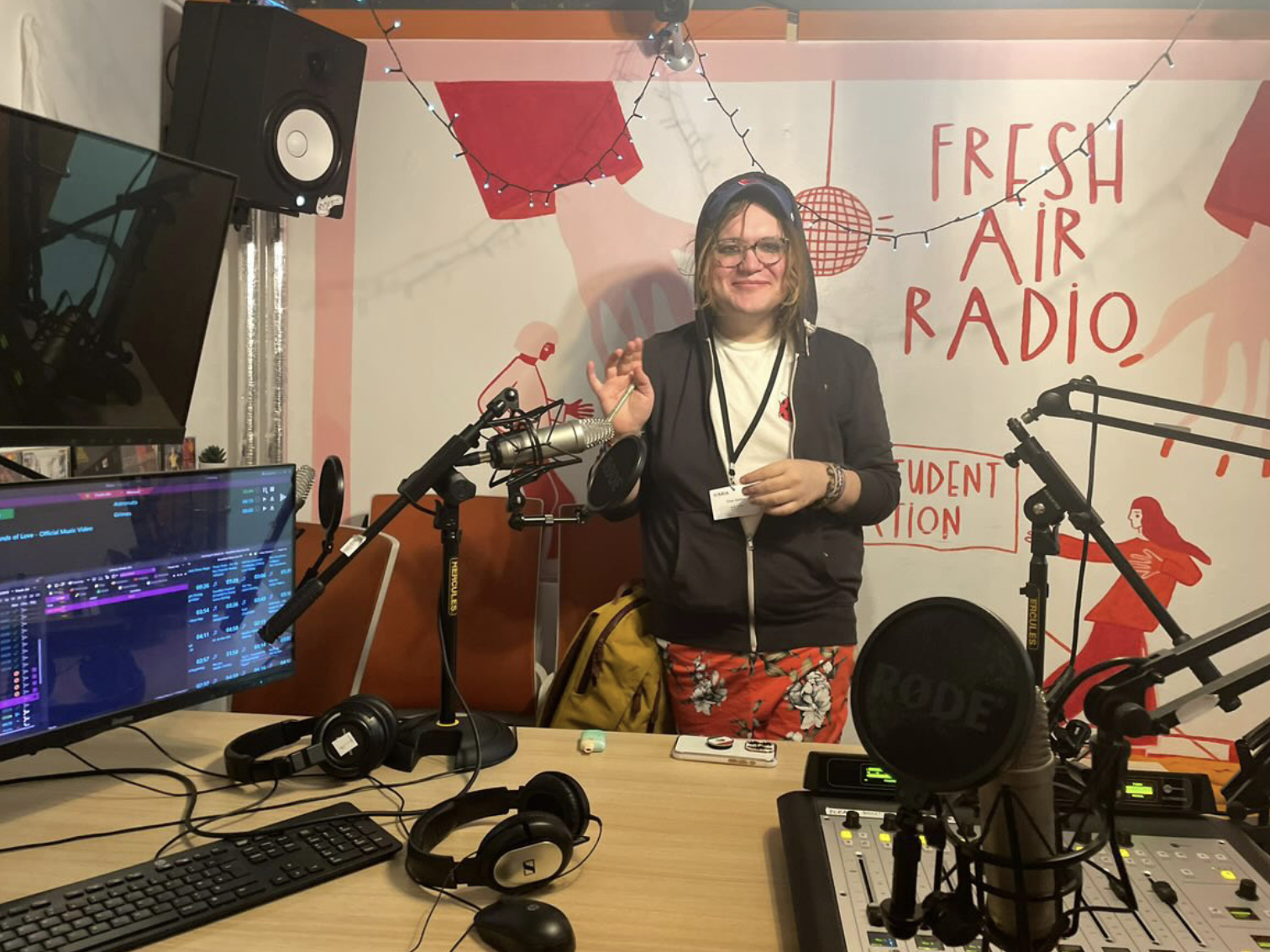 chee waving at cam standing in radio show with fresh air radio on wall