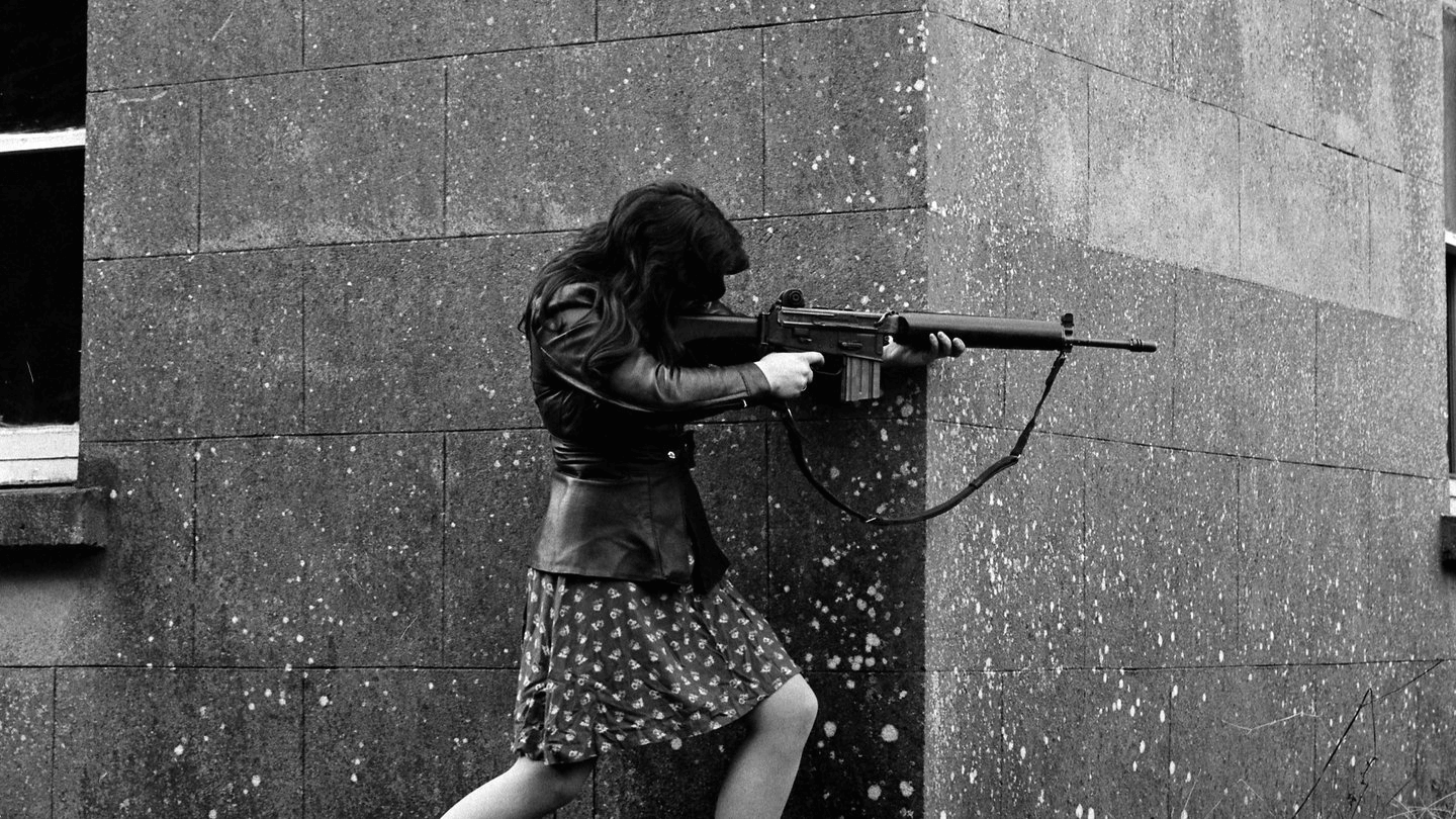 woman in cute dress and leather jacket pointing an AR-18 assault rifle around
a corner