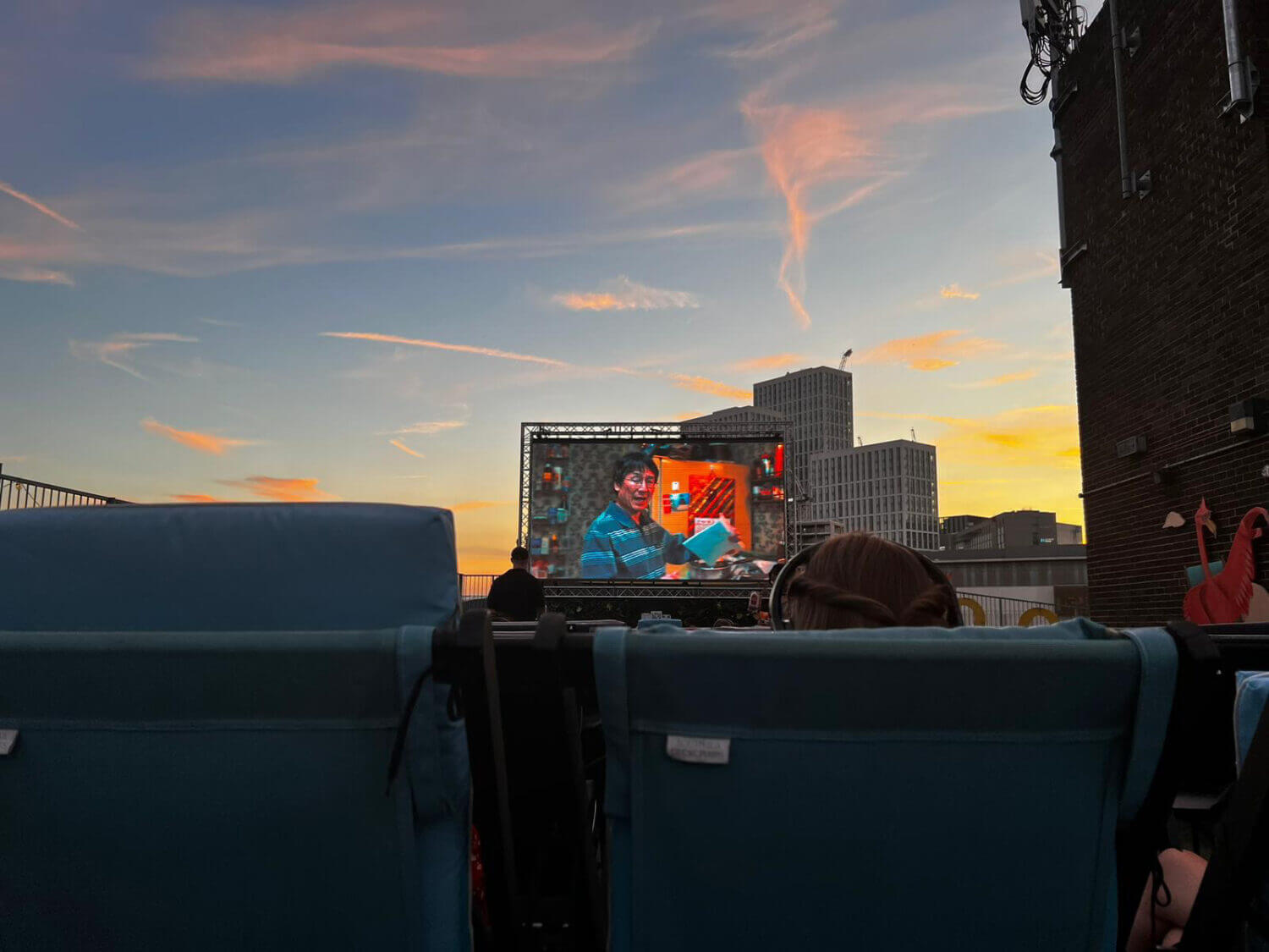 photo of sunset from rooftop. cinema screen in center of photo. chairs block off the lower half of the photo. the sky is pink and purple, gold and grey.