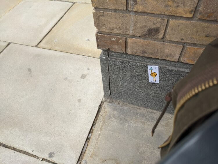 a photo of a sticker on a marble wall. the wall is a few feet away, and the floor and a coat are visible, the sticker is near the bottom of the wall. the sticker depicts a giraffe with a green triangle head and fancy heeled shoes