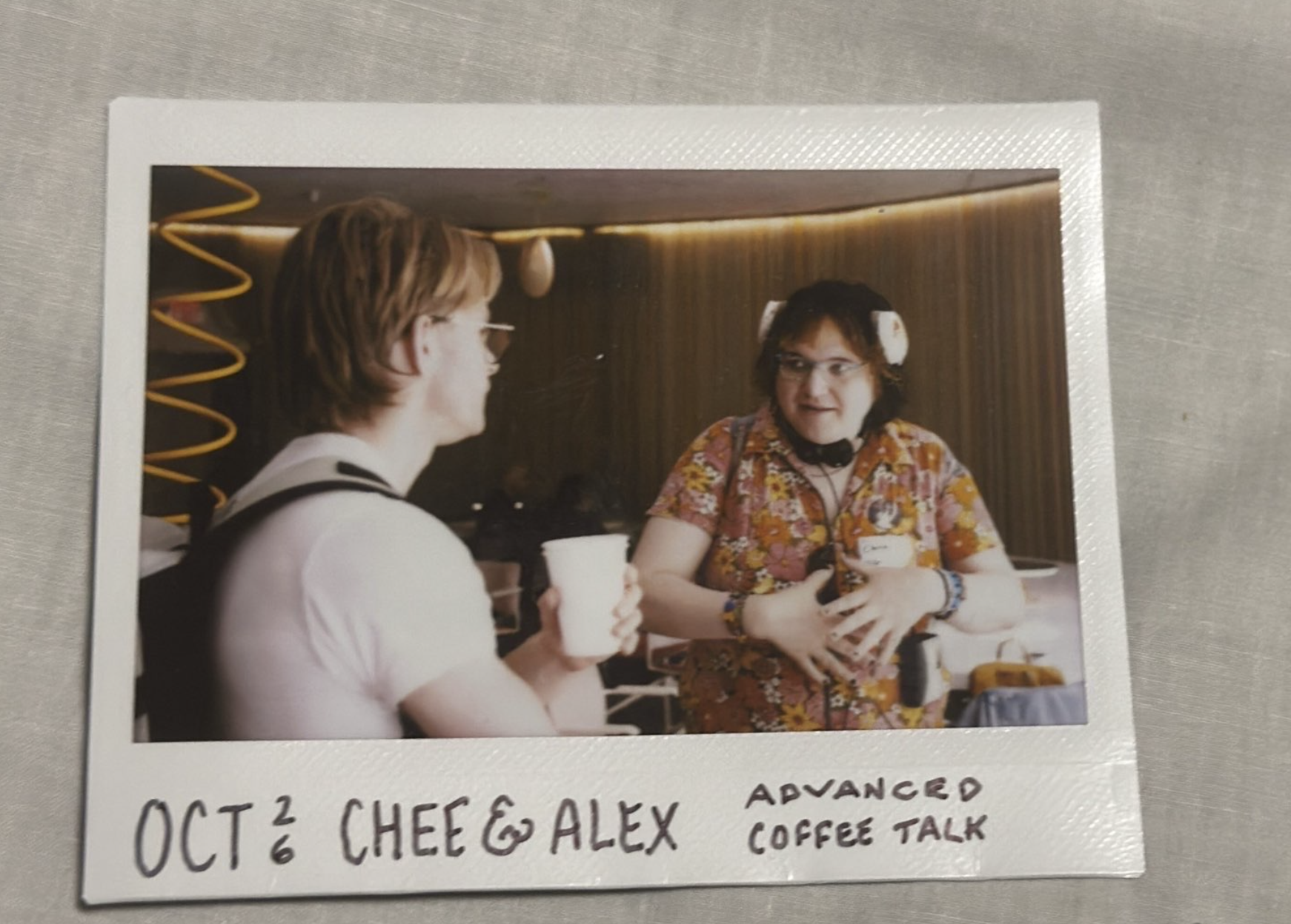 a photo of an instax printed photo of me in a flowery jumpsuit gesticulating
wildly. i have a hungry look on my face and am in the middle of a word. i have a
pair of headphones on my head, and another around my neck. alex good is next to
me holding a coffee and humouring me with a convincing job of paying
attention